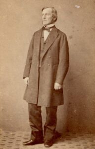 Sepia coloured studio photograph of George Boole in a long-coat suit with dickie-bow tie standing with his hands at his side, his head slightly turned to the left as we look at it.