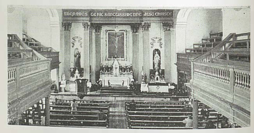 An image of the old St Francis Church in Cork City with pews in front of the altar. There are statues on either side of the altar and Latin writing over the altar and statues.