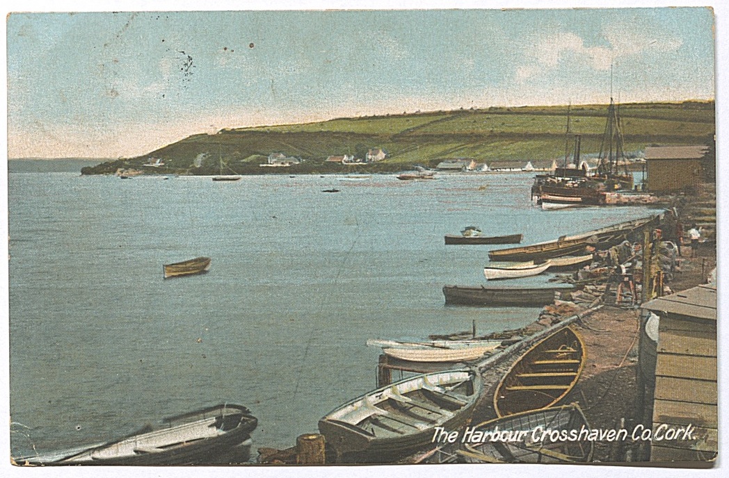 Title in lower right: The Harbour Crosshaven, Co Cork. It's a historical view of Crosshaven harbour. On the right are lots of small boats and sailing ships tied up at the dock or berthed. A few small rowing boats are anchored in the sea. The sea and harbour are in the centre of the image. In the background are various houses on a peninsula. 