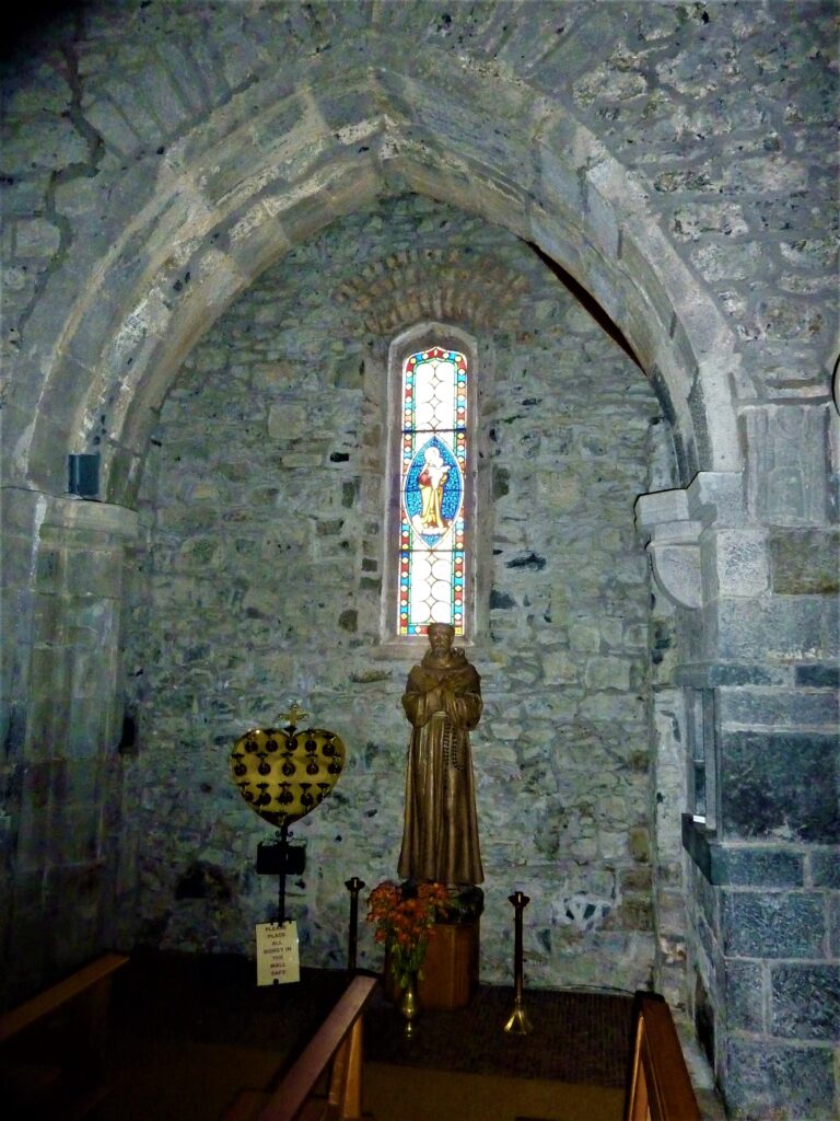 An archway opening off the south chapel in Meelick friary. There is a statue of St Francis, candles and pews visible. The surrounds of the friary walls are stone.