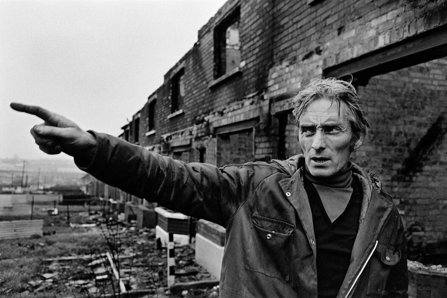 Black and white photograph by Alen MacWeeney of man with outstretched right arm. Ruins in background. Belfast, 1971. 
