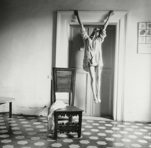 Black and white photography by Francesca Woodman of figure in light shirt hanging from a door frame. Self-portrait.