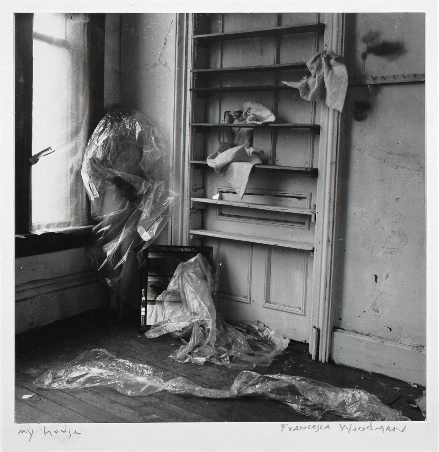Black and white photograph by Francesca Woodman of figure standing in a corner covered in plastic. Self-portrait.