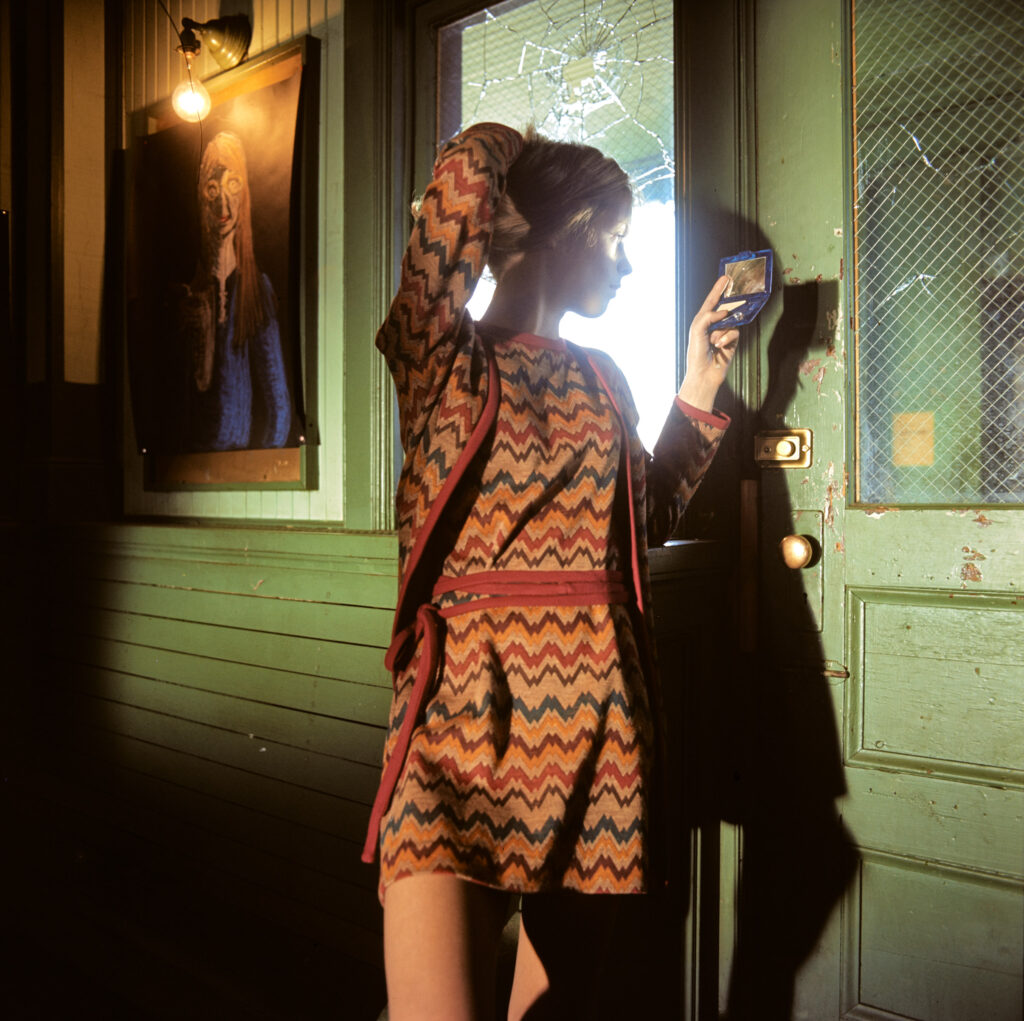 Colour fashion photograph of woman looking out of a window and holding a compact mirror in one hand. by Alen MacWeeney. This is an example of fashion photography.