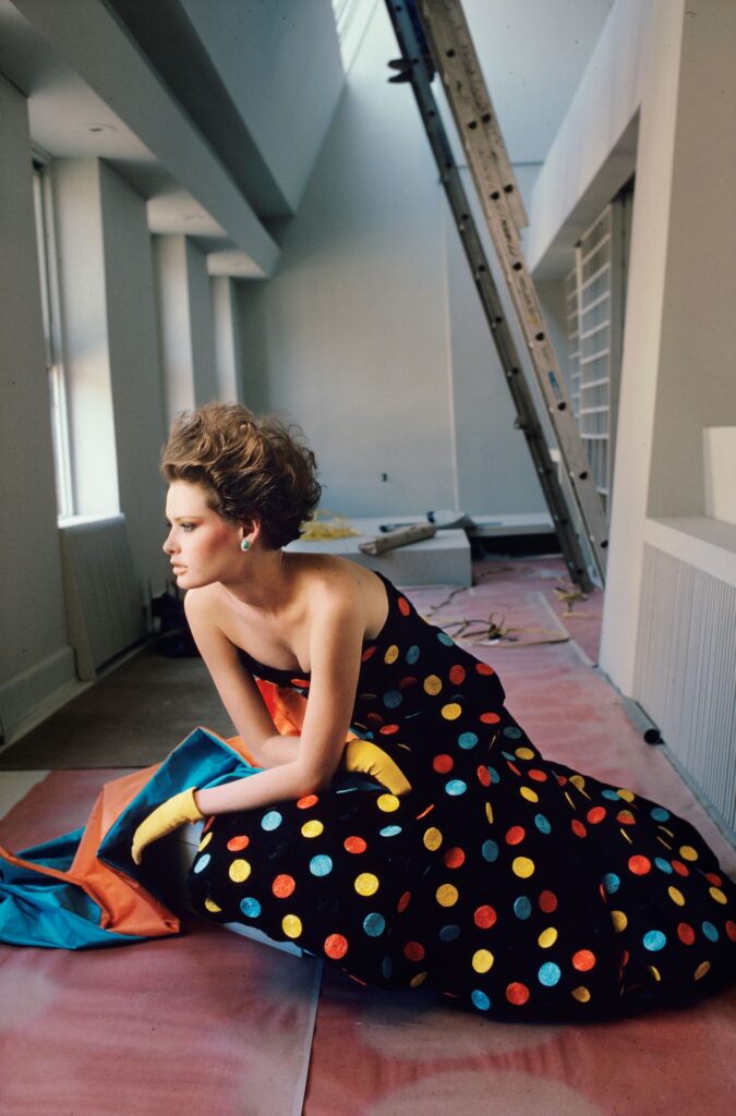 Colour fashion photograph by Alen MacWeeney. Woman sitting on floor wearing black gown with coloured dots. Ladder in background. Photographed for designer Arnold Scassi. This is an example of fashion photography.