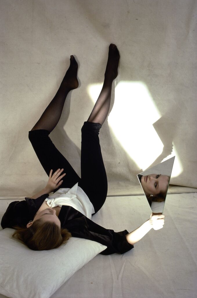 Colour fashion photo by Alen MacWeeney. Woman lying on floor with legs up against the wall. Model is holding mirror in right hand, face reflected in mirror. This is an example of fashion photography.