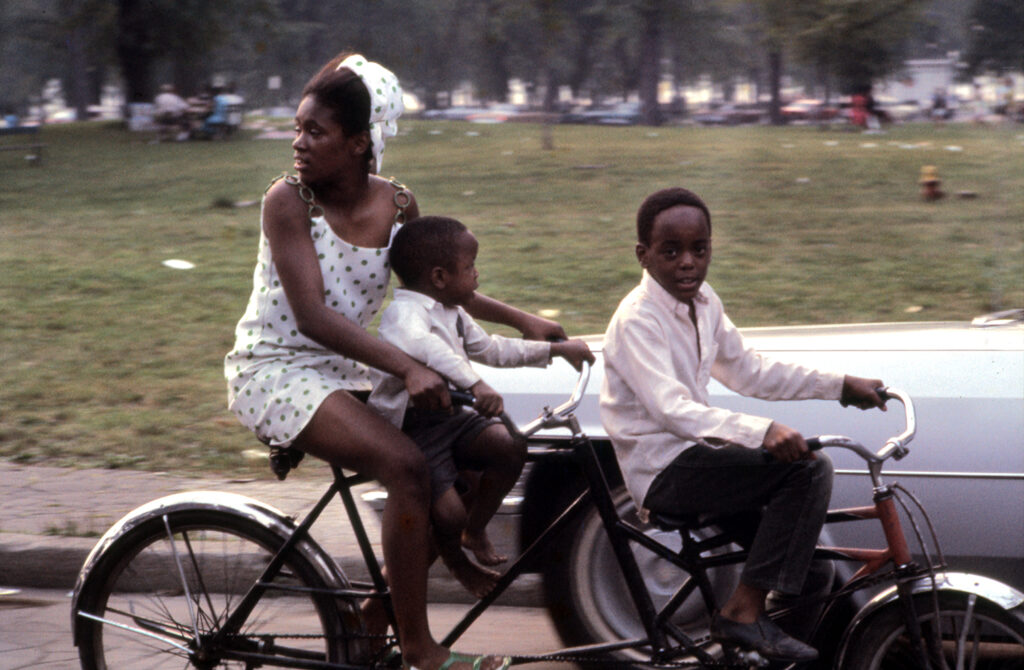 One African-American woman and two children riding on two bicycles. In Detroit, Michigan. Colour photography by Alen MacWeeney. Black History Month.