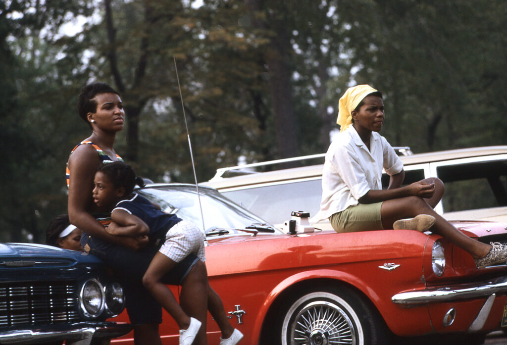 Two African-American women and one child sitting on the hoods of two cars. In Detroit Michigan. Colour photo by Alen MacWeeney. Black History Month.