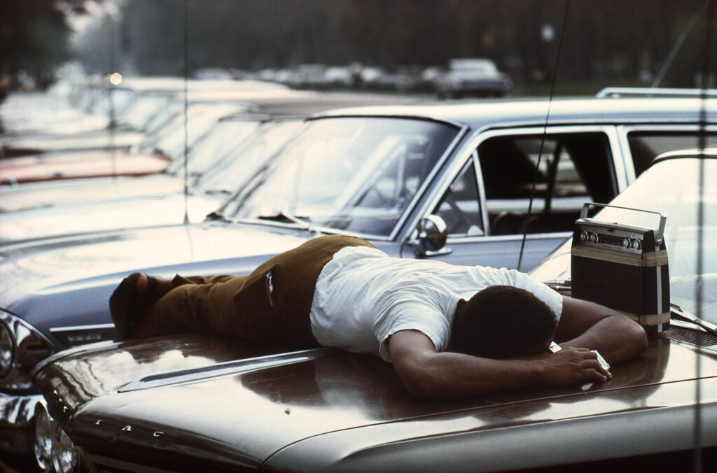 African-American man lying face down on a car hood in Detroit, Michigan. Colour photo by Alen MacWeeney. Black History Month.