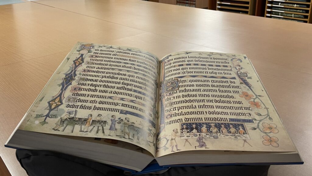 Opening in the facsimile of the Luttrell Psalter. The text is in Latin with many decorations surrounding the text. At the foot of the folio on the right are various people feasting.