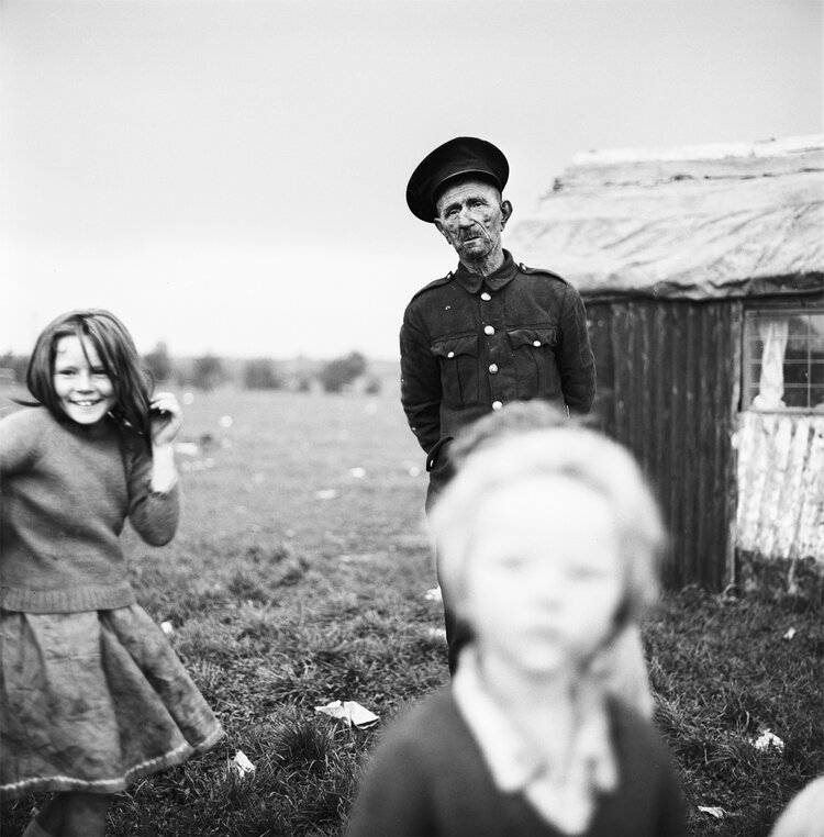 Black and white photo of man with two children, one in foreground blurred, the other to the left. Taken by Alen MacWeeney.