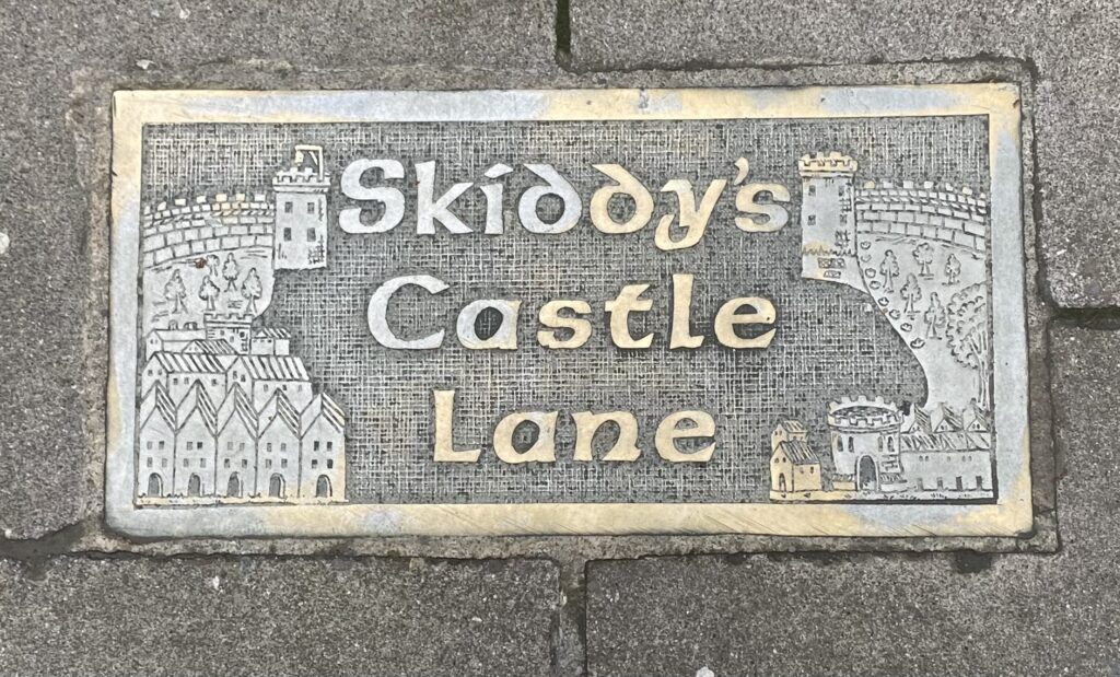 Photograph of the plaque for Skiddy's Castle Lane in the footpath. It gives the name between two towers and various houses and trees.