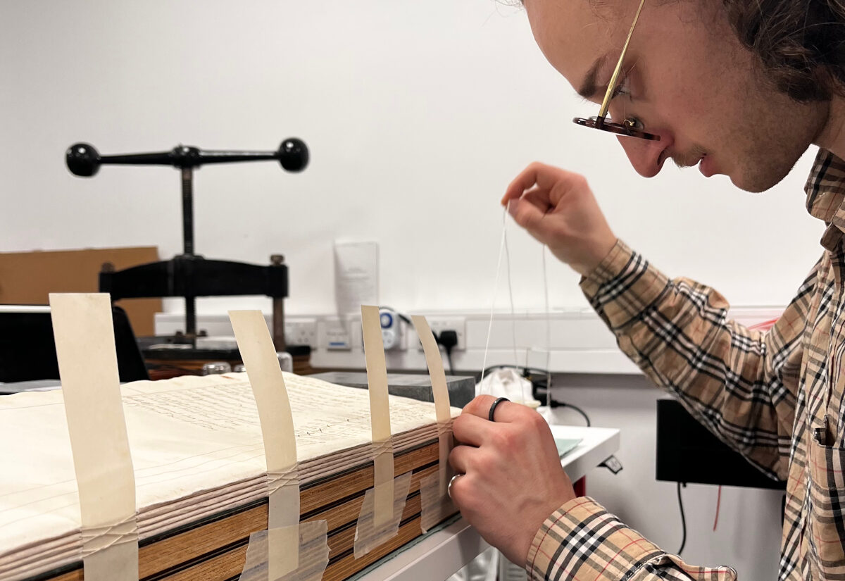 conservation intern pulls thread through the spine of the ledger, to sew it together.
