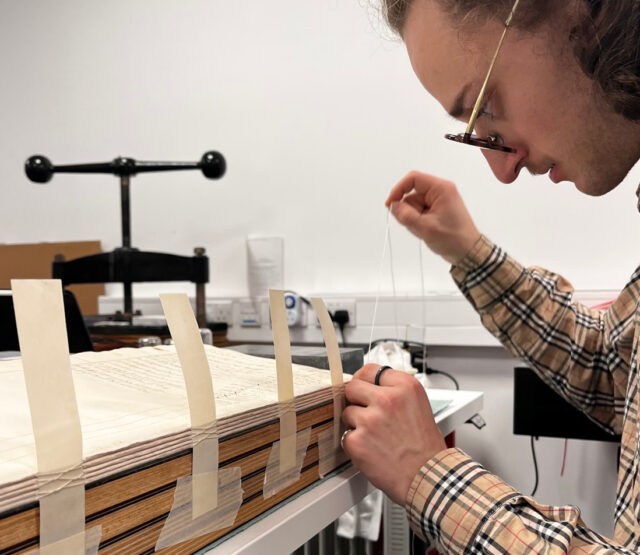 conservation intern pulls thread through the spine of the ledger, to sew it together.