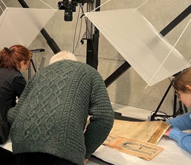 Three people examine a manuscript. Their backs are to the camera.